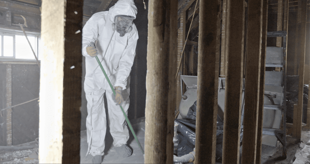 A Foralis Environmental professional asbestos abatement worker wearing full protective gear and a respirator is carefully cleaning up asbestos debris inside a stripped-down building, ensuring safe removal and compliance with WorkSafeBC regulations.